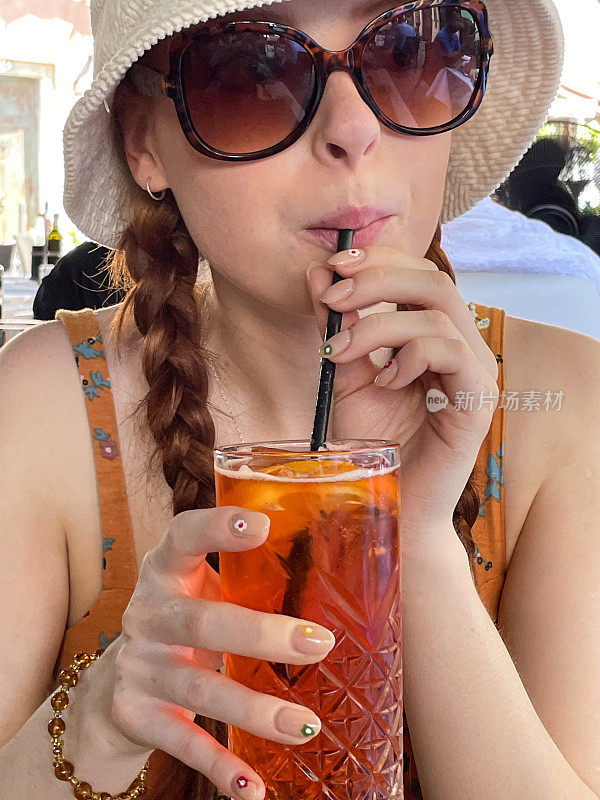 全画幅图片，迷人的红头发的女人喝着Spritz Veneziano鸡尾酒饮料与塑料吸管，冰块和橙子片，户外戴着太阳镜，遮阳帽和太阳裙与穆拉诺玻璃手镯，户外用餐设置背景